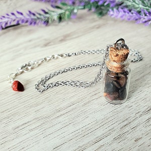 May include: A silver chain necklace with a small glass bottle pendant filled with brown, black, and red stones. The bottle has a cork stopper and is attached to the chain with a silver loop.