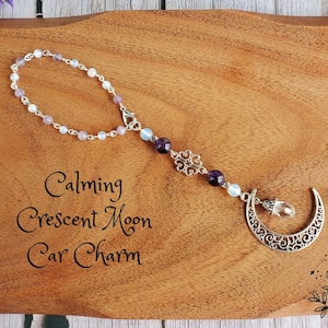May include: A silver crescent moon car charm with a beaded chain featuring amethyst and opal-like beads. The charm is on a wooden surface with the text "Calming Crescent Moon Car Charm".