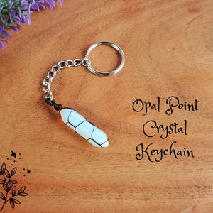 May include: A crystal keychain with a silver chain and ring. The crystal is a light blue color with a black web-like pattern. The text "Opal Point Crystal Keychain" is written in black script.