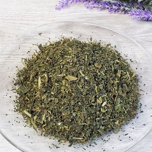 May include: A close-up of a bowl filled with dried green herbs. The herbs are a mix of different types, including leaves, stems, and flowers.