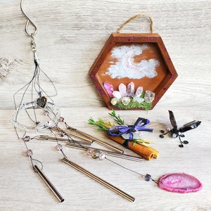 May include: A crystal and gemstone wind chime with a wooden hexagon frame featuring a crescent moon and clouds. The chime has a silver heart charm and pink and purple crystals. The frame is decorated with moss and small crystals.