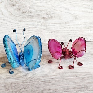 May include: Two wire-wrapped butterflies made with agate slices. One butterfly is blue and the other is pink.