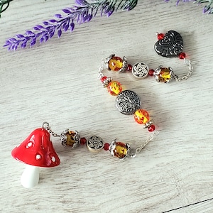 May include: A whimsical necklace featuring a red mushroom charm with white spots, connected to a chain of amber-colored beads, silver-toned decorative beads, and small red beads. A silver heart charm completes the design. The necklace rests on a light wood surface.