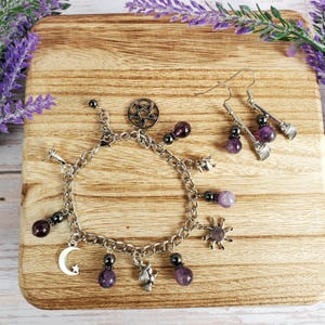 May include: A silver charm bracelet and matching earrings with a witchy theme. The bracelet features charms like a crescent moon, sun, and pentagram, along with purple gemstone beads. The earrings have broomstick charms and purple beads. The jewelry is displayed on a wooden surface.