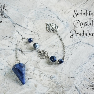 May include: A sodalite crystal pendulum with a silver chain and decorative accents. The pendulum features a blue, cone-shaped crystal and is adorned with blue and gray beads. The text "Sodalite Crystal Pendulum" is written in script.