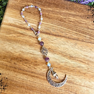 May include: A silver-toned car charm featuring a crescent moon pendant with a clear crystal and a beaded chain. The chain includes purple and white beads, and a decorative silver charm. The charm is displayed on a wooden surface.