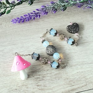May include: A pink mushroom charm with a silver chain attached to a beaded necklace with blue and pink beads and silver charms. The necklace has a silver heart charm with a floral design.