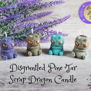 May include: Four small, hand-painted dragon candles in purple, green, blue, and gray. Each candle has gold eyes and a grumpy expression. The candles are arranged on a wooden surface with lavender sprigs in the background. The text "Disgruntled Pine Tar Scrap Dragon Candle" is written below the candles.