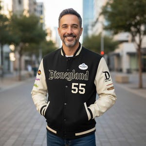 Może przedstawiać: Czarna i kremowa kurtka typu letterman z napisem "Disneyland 55" w białym skrypcie. Kurtka ma kremowe rękawy, czarny korpus i biały kołnierz. Na rękawach widoczne są naszywki.