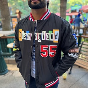 May include: Black bomber jacket with a colorful "Disneyland" patch on the front and a red "55" on the chest. The jacket has a white and red striped collar and cuffs.