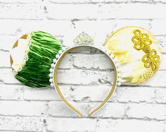 Può includere: Una fascia decorativa con due accenti rotondi a forma di orecchio. Un orecchio è verde con un motivo strutturato, l'altro è bianco e oro con ricami floreali dorati. La fascia ha una tiara argentata e dettagli di perle bianche.