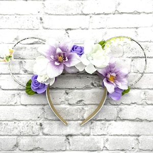 May include: Purple and white flower headband with a silver wire frame and a small fairy figurine. The headband is on a white brick background.