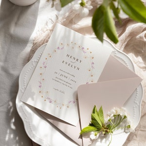 Puede incluir: Una invitaci&oacute;n de boda con un dise&ntilde;o de corona floral. La invitaci&oacute;n es para la boda de Henry y Evelyn el 30 de junio de 2025. La invitaci&oacute;n est&aacute; sobre un fondo blanco con un sobre rosa.