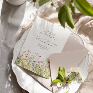 May include: A wedding invitation with a floral design featuring a watercolor illustration of wildflowers. The invitation is printed on white cardstock and has a pink envelope. The text on the invitation reads "Chris & Rosie invite you to their wedding Saturday, May 25, 2030 Four o'clock in the afternoon 123 Anywhere st. Any City, St 12345"