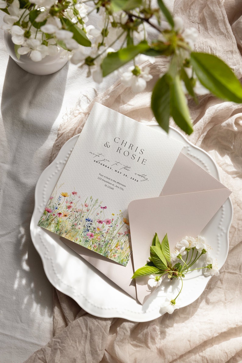 May include: A wedding invitation with a floral design featuring a watercolor illustration of wildflowers. The invitation is printed on white cardstock and has a pink envelope. The text on the invitation reads "Chris & Rosie invite you to their wedding Saturday, May 25, 2030 Four o'clock in the afternoon 123 Anywhere st. Any City, St 12345"