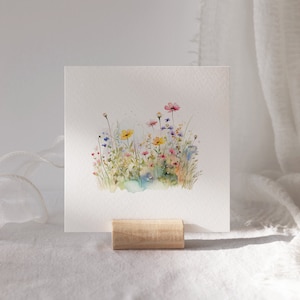 May include: A watercolor painting of a field of wildflowers in shades of pink, yellow, blue, and green. The flowers are arranged in a cluster, with some stems extending beyond the group. The painting is on a white background.