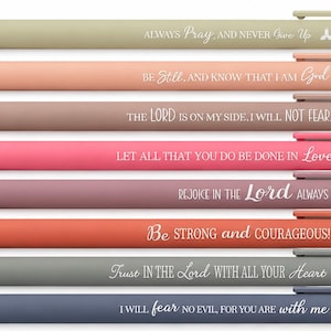 May include: A set of eight retractable ballpoint pens in various muted colors, each featuring a unique white printed religious quote and a small icon. The pens are arranged horizontally, displaying phrases like Always Pray, Be Still, and Trust in the Lord.