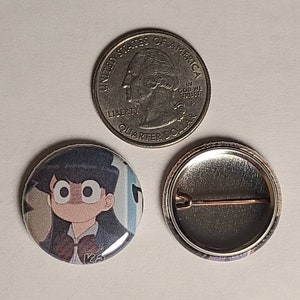 May include: A round button with a cartoon image of a girl with black hair and large eyes. The girl is wearing a school uniform with a bow tie. The button is 1 inch in diameter and has a silver metal back.