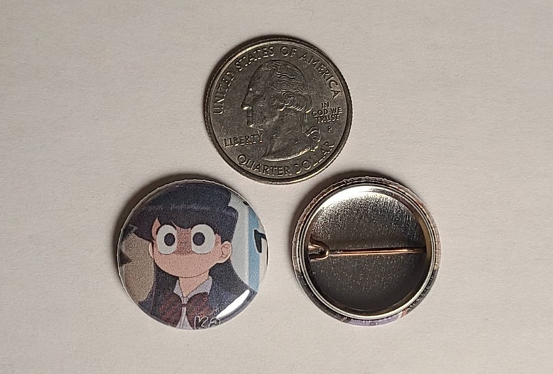 May include: A round button with a cartoon image of a girl with black hair and large eyes. The girl is wearing a school uniform with a bow tie. The button is 1 inch in diameter and has a silver metal back.