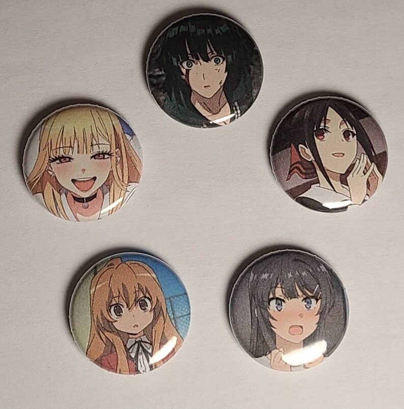 May include: Five round button pins featuring anime characters. The pins are colorful and feature different characters with various expressions.