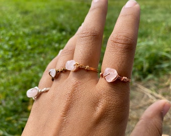 May include: Three rings with pink rose quartz stones wrapped in gold wire. The rings are worn on a person's hand.