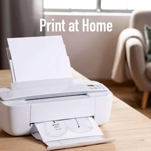 May include: A white inkjet printer on a wooden table, printing a sheet of paper with a mask pattern. The text "Print at Home" is displayed above the printer. A sofa and window are visible in the background.