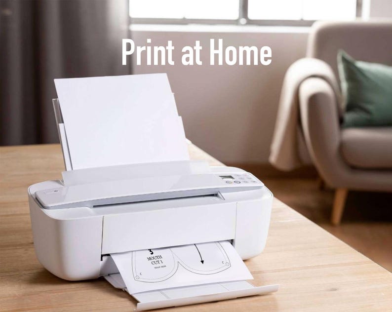 May include: A white inkjet printer on a wooden table, printing a sheet of paper with a mask pattern. The text "Print at Home" is displayed above the printer. A sofa and window are visible in the background.