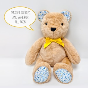May include: A plush brown teddy bear with a yellow bow, blue and white floral ears and feet. The bear is sitting on a white surface. The text "I'm soft, cuddly, and safe for all ages!" is in a speech bubble above the bear.