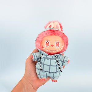 May include: A plush toy with a round face, red eyes, and a toothy grin. It wears a blue plaid hoodie with a pink fur-lined hood and bunny ears. The toy is held against a light blue background.