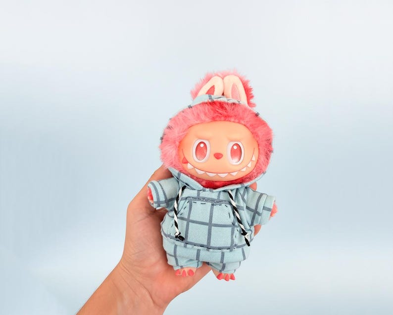 May include: A plush toy with a round face, red eyes, and a toothy grin. It wears a blue plaid hoodie with a pink fur-lined hood and bunny ears. The toy is held against a light blue background.