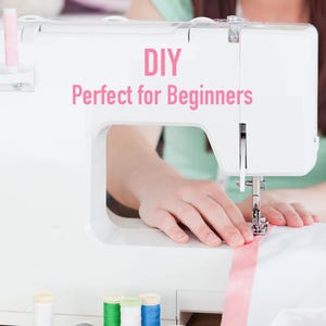 May include: A white sewing machine with pink thread spools and the text "DIY Perfect for Beginners." A person is sewing a pink ribbon onto white fabric. The machine has a needle and presser foot.