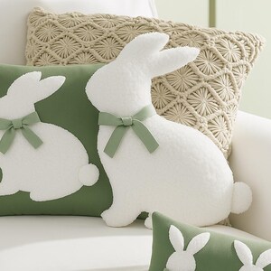 May include: Two plush white bunny pillows with green bows. One bunny pillow is on a green pillow with a white bunny silhouette. The other bunny pillow is on a beige crocheted pillow.