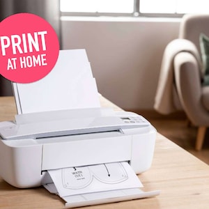 May include: A white printer with a sheet of paper feeding through it. The paper has a pattern for a mouth mask printed on it. The text "PRINT AT HOME" is on a pink circle in the upper left corner of the image.