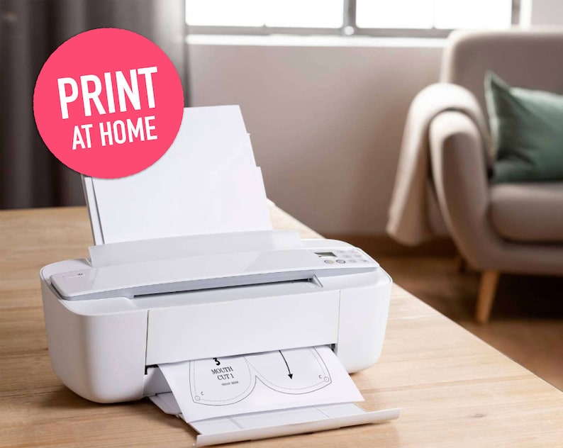 May include: A white printer with a sheet of paper feeding through it. The paper has a pattern for a mouth mask printed on it. The text "PRINT AT HOME" is on a pink circle in the upper left corner of the image.