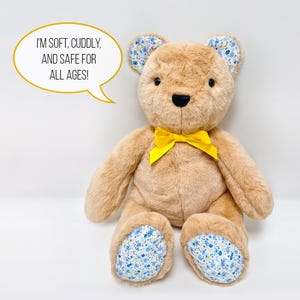 Pu&ograve; includere: Un orsacchiotto di peluche marrone con un fiocco giallo, orecchie e piedi con fiori blu e bianchi. L'orso &egrave; seduto su una superficie bianca. Il testo "Sono morbido, coccoloso e sicuro per tutte le et&agrave;!" &egrave; in un fumetto sopra l'orso.