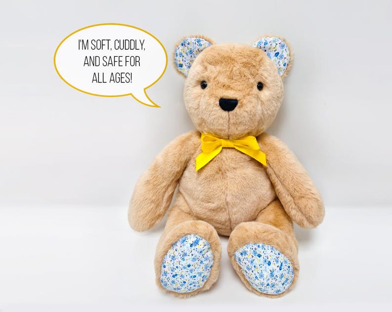 Pu&ograve; includere: Un orsacchiotto di peluche marrone con un fiocco giallo, orecchie e piedi con fiori blu e bianchi. L'orso &egrave; seduto su una superficie bianca. Il testo "Sono morbido, coccoloso e sicuro per tutte le et&agrave;!" &egrave; in un fumetto sopra l'orso.