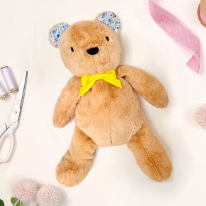 May include: A brown teddy bear with blue floral fabric ears and a yellow bow tie. The bear is lying on a white surface with sewing supplies nearby.