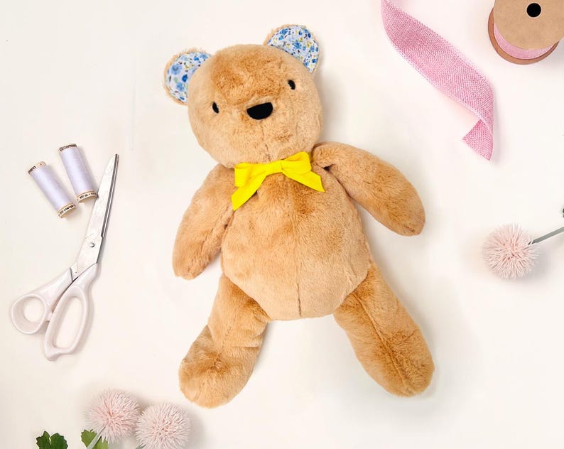 May include: A brown teddy bear with blue floral fabric ears and a yellow bow tie. The bear is lying on a white surface with sewing supplies nearby.