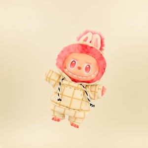 May include: A plush toy figure with a smiling face, red eyes, and a pink furry hood with bunny ears. The figure wears a cream-colored plaid jumpsuit with a drawstring hood and black accents. The toy has small arms and legs.