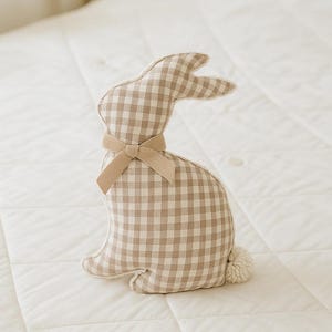 May include: A beige and brown gingham bunny rabbit plush toy with a brown velvet bow. The bunny is sitting on a white quilted surface.