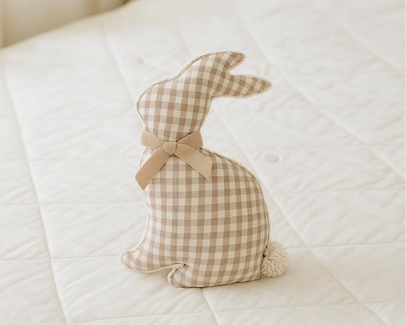 May include: A beige and brown gingham bunny rabbit plush toy with a brown velvet bow. The bunny is sitting on a white quilted surface.