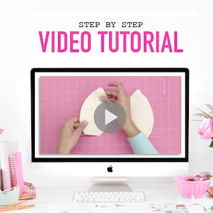 May include: A computer screen displaying a video tutorial on how to make a craft project. The video shows a person's hands holding a piece of white fabric on a pink cutting mat. The video is titled "Step by Step Video Tutorial".