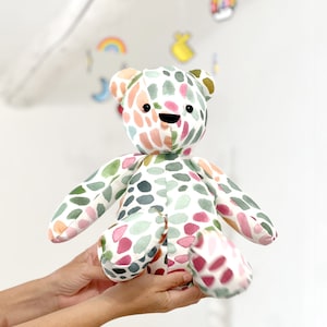 May include: A colorful, hand-painted teddy bear with a white background and green, pink, and orange watercolor spots. The bear has black eyes and a black smile.