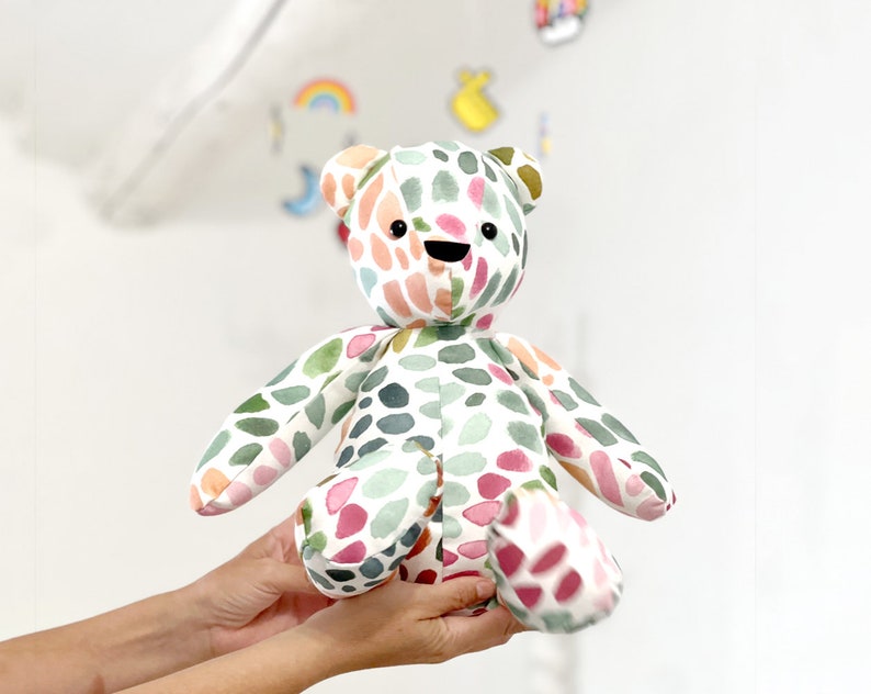 May include: A colorful, hand-painted teddy bear with a white background and green, pink, and orange watercolor spots. The bear has black eyes and a black smile.