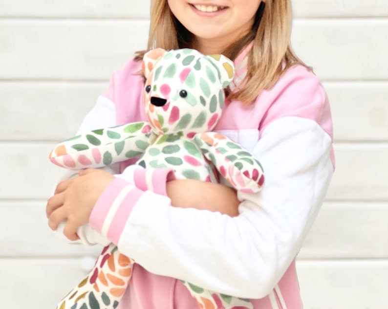 May include: A child is holding a colorful patchwork teddy bear with green, pink, and yellow accents. The bear has black button eyes and a black stitched nose.