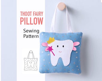 Puede incluir: Una almohada azul para el hada de los dientes con un diseño de diente blanco, una corona dorada y una varita rosa. La almohada tiene un asa blanca y el texto "THOOT FAIRY PILLOW Sewing Pattern".