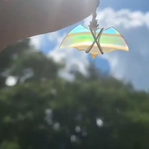 May include: A small, iridescent bat-shaped pendant, suspended by a silver-colored wire. The pendant reflects a spectrum of colors, including yellow, blue, and green. The background features a blurred view of green foliage and a bright, sunny sky.