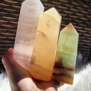 May include: Three polished crystal towers in shades of pink, yellow, and green are held in a hand. The crystals have a smooth, faceted shape and display natural variations in color and pattern. The background is a woven texture.