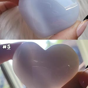 May include: Two heart-shaped stones, one light blue and the other a pale lavender, are held in a person's hand. The stones are smooth and polished, with a translucent quality. The text "#5" is visible on the bottom stone. These are likely decorative or collectible items.