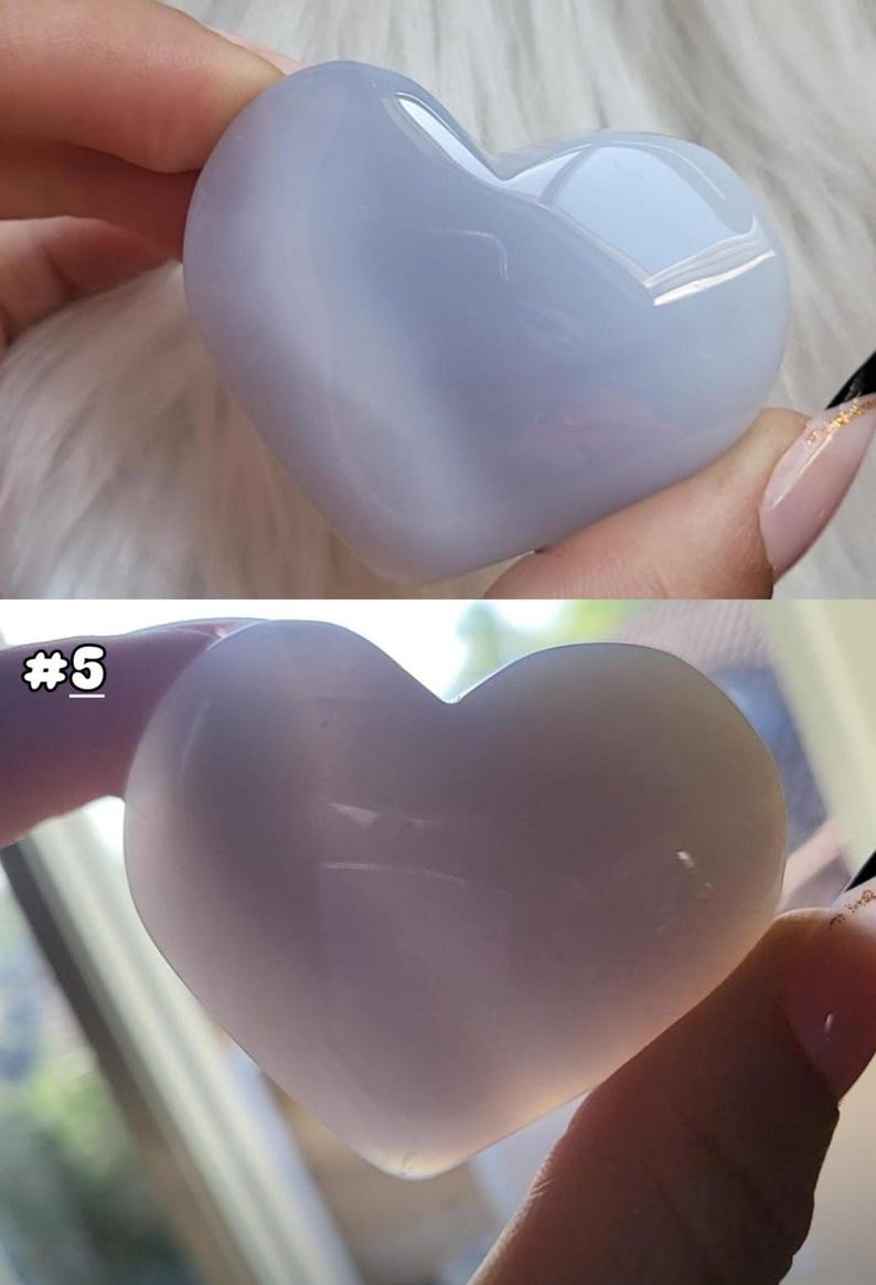 May include: Two heart-shaped stones, one light blue and the other a pale lavender, are held in a person's hand. The stones are smooth and polished, with a translucent quality. The text "#5" is visible on the bottom stone. These are likely decorative or collectible items.